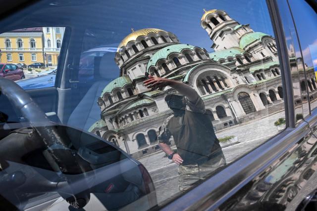 A reflection in a car window shows a masked soldier of the Ukrainian army waving to a fellow soldier, during a ceremony to hand over 17 off-road vehicles donated from Bulgarians and Russians, living in Bulgaria, in front of Alexander Nevsky Cathedral in Sofia on July 1, 2026. (Photo by Nikolay DOYCHINOV / AFP)