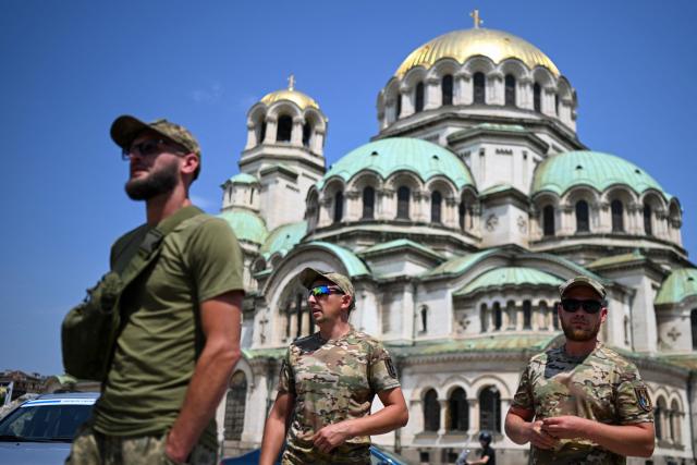 Soldiers of the Ukrainian army with Ukrainian flags walk next to donated cars during a ceremony to hand over 17 off-road vehicles donated by Bulgarians and Russians living in Bulgaria, in front of the Alexander Nevsky Cathedral in Sofia on July 1, 2026. (Photo by Nikolay DOYCHINOV / AFP)