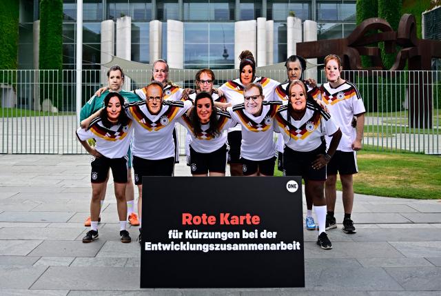 Members of global development aid organization ONE pose with masks with the faces of German cabinet ministers and with the jerseys of the German national football team during a protest against massive cuts in development cooperation under the motto "Red Card for Merz" in Berlin on July 1, 2026. The activists wear masks with the faces of (front row, LtoR) German Minister for Economic Cooperation and Development Reem Alabali-Radovan, German Chancellor Friedrich Merz, German Minister for Research, Technology and Aerospace Dorothee Baer, German Foreign Minister Johann Wadephul, German Minister for Labour and Social Affairs Baerbel Bas, and (back row, LtoR) German Finance Minister and Vice Chancellor Lars Klingbeil, German Defence Minister Boris Pistorius, German Interior Minister Alexander Dobrindt, German Economy and Energy Minister Katherina Reiche, Chief of Staff and Minister for Special Tasks Thorsten Frei and German Minister for Education, Family, Senior Citizens, Women and Youth Karin Prien. The federal government is set to adopt its draft budget for 2027 next week. (Photo by Tobias SCHWARZ / AFP)