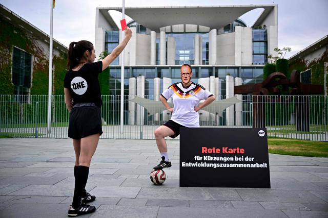 A member of global development aid organization ONE posing with a mask with the face of German Chancellor Friedrich Merz and wearing a jersey of the German national football team is shown a red card during a protest against massive cuts in development cooperation under the motto "Red Card for Merz" in Berlin on July 1, 2026. The federal government is set to adopt its draft budget for 2027 next week. (Photo by Tobias SCHWARZ / AFP)