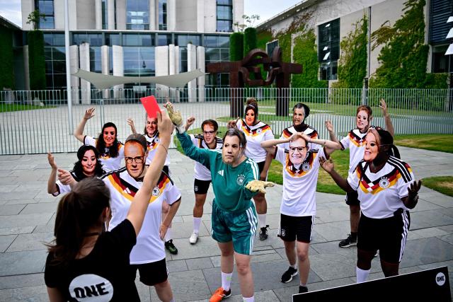 Members of global development aid organization ONE with masks with the faces of German Chancellor Friedrich Merz (3rdL) and German cabinet ministers and wearing the jerseys of the German national football team are shown the red card during a protest against massive cuts in development cooperation under the motto "Red Card for Merz" in Berlin on July 1, 2026. The federal government is set to adopt its draft budget for 2027 next week. (Photo by Tobias SCHWARZ / AFP)