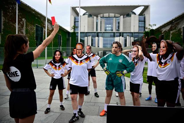 Members of global development aid organization ONE with masks with the faces of German Chancellor Friedrich Merz (3rdL) and German cabinet ministers and wearing the jerseys of the German national football team are shown the red card during a protest against massive cuts in development cooperation under the motto "Red Card for Merz" in Berlin on July 1, 2026. The federal government is set to adopt its draft budget for 2027 next week. (Photo by Tobias SCHWARZ / AFP)