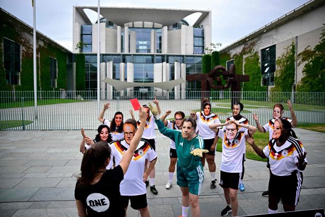 Members of global development aid organization ONE with masks with the faces of German Chancellor Friedrich Merz (3rdL) and German cabinet ministers and wearing the jerseys of the German national football team are shown the red card during a protest against massive cuts in development cooperation under the motto "Red Card for Merz" in Berlin on July 1, 2026. The federal government is set to adopt its draft budget for 2027 next week. (Photo by Tobias SCHWARZ / AFP)