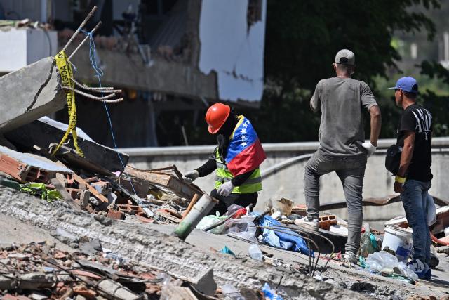A man wrapped in a Venezuelan national flag tries to find missing people amid the rubble at Los Cocos beach, in Caraballeda, La Guaira state, Venezuela, on July 1, 2026, following the June 24 twin earthquakes. Venezuelans, with help from around the world, were still searching on July 1, 2026 for survivors a week after massive earthquakes killed nearly 2,000 and left thousands unaccounted for. (Photo by Juan BARRETO / AFP)