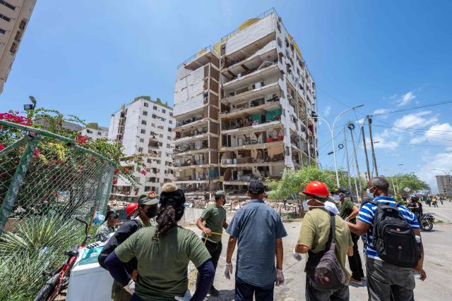 People stand near a damaged building in Playa Grande neighborhood, La Guaira state, Venezuela, on July 1, 2026, following the June 24 twin earthquakes. Tens of thousands of people urgently need food and shelter in Venezuela due to the devastation from two huge earthquakes that killed more than 1,700 and injured 5,000, the UN said on June 30, 2026, as doctors warned of potential outbreaks of disease. (Photo by Miguel MEDINA / POOL / AFP)