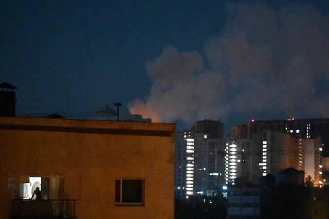 A man (front L) looks out from his flat in a residential building as smoke rises in the background during a Russian missile strike on the Ukrainian capital Kyiv on July 2, 2026, amid the Russian invasion in Ukraine. Russian missile and drone strikes rocked Kyiv early on July 2, setting off fires and wounding at least five people, after President Volodymyr Zelensky warned that Moscow was preparing a "massive attack". (Photo by Sergei SUPINSKY / AFP)