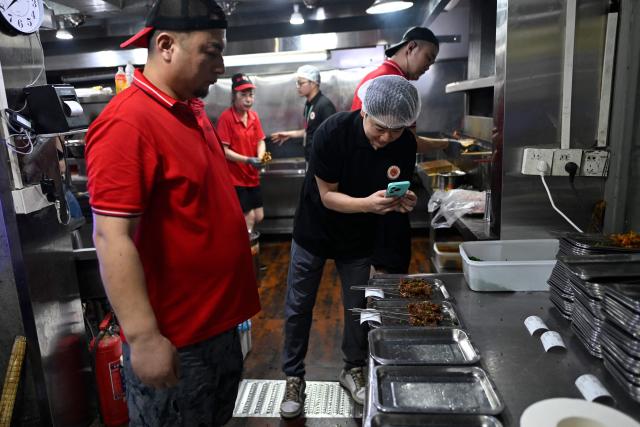 This picture taken on June 15, 2026 shows a trainee using a mobile phone to take pictures during a visit of a barbecue restaurant in Yueyang, in southern China's Hunan province. Hunched over sizzling grills and shrouded in smoke drifting from white-hot charcoal, dozens of trainees at the Yueyang Barbecue Academy in southern China roast skewer after skewer of succulent beef tendon to perfect the fragrant, spicy taste of their homework. (Photo by WANG Zhao / AFP) / TO GO WITH: China-culture-food-bbq, by Sam DAVIES