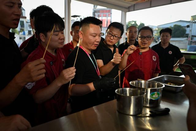 This picture taken on June 15, 2026 shows a trainer (C with a lanyard) and a group of trainees tasting skewers during practical session at the Yueyang Barbecue Academy in Yueyang, in southern China's Hunan province. Hunched over sizzling grills and shrouded in smoke drifting from white-hot charcoal, dozens of trainees at the Yueyang Barbecue Academy in southern China roast skewer after skewer of succulent beef tendon to perfect the fragrant, spicy taste of their homework. (Photo by WANG Zhao / AFP) / TO GO WITH: China-culture-food-bbq, by Sam DAVIES