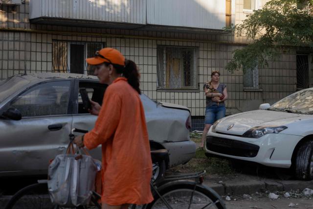 A woman holds a dog next to damaged cars as another passes by with a bicycle and a smartphone following a Russian air attack on the Ukrainian capital of Kyiv on July 2, 2026, amid the Russian invasion in Ukraine. Russia launched its largest ever barrage on Kyiv early July 2, 2026, according to the city's mayor, tearing open apartment buildings in an hours-long drone and missile attack that killed at least 21 people. (Photo by Tetiana DZHAFAROVA / AFP)