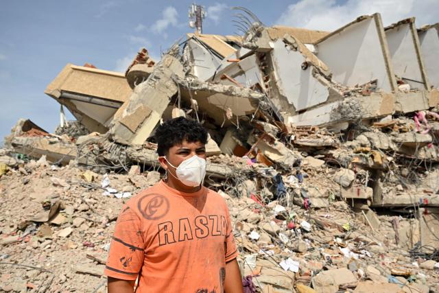 Miguel Cordoba, 20, who survived after escaping from his building (behind) collapsed during twin earthquakes, walks amid the rubble in Caraballeda, La Guaira state, Venezuela, on July 2, 2026. Hundreds of rescuers in Venezuela cheered and embraced on July 2, 2026 after pulling 43-year-old Hernan Gil from the ruins of a collapsed building eight days after deadly twin earthquakes, AFP journalists witnessed. (Photo by MARTIN BERNETTI / AFP)