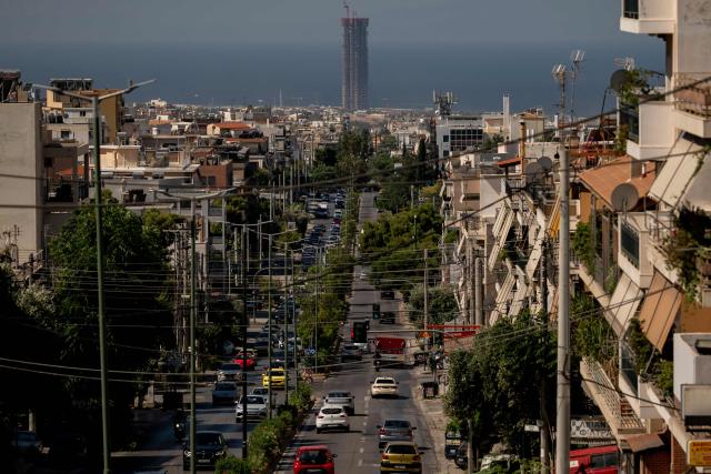 People drive their vehicles on a main road of an Athenian suburb on June 26, 2026, as a 200-meter-tall, 50-story skyscraper towers which is is the tallest building ever constructed in Athens is seen in the background. Less than 15 km from the center of Athens, Glyfada—like many neighborhoods along the Saronic Gulf—has seen its population grow in recent years due to a major real estate development underway in the neighboring municipality of Ellinikon, leading to skyrocketing prices.
Nicknamed “The Athenian Riviera”, this massive development project, spanning an area of 6.2 kmІ, will soon include hotels, shopping centers, parks, marinas, 8,000 housing units, and a “new city” of 30,000 residents, according to Lamda Development, the company in charge. (Photo by Aris MESSINIS / AFP)