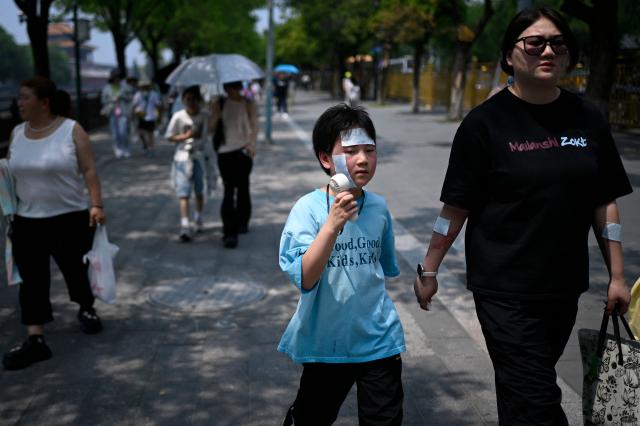 A child, holding a mini fan with cooling patches stuck to his face, walks down a road with his family near the Forbidden City on a hot day in Beijing on July 3, 2026. (Photo by WANG Zhao / AFP)