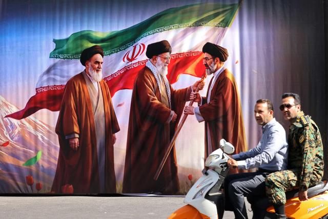 Men ride a scooter past a billboard featuring the images of the late founder of the Islamic Revolution supreme leader Ayatollah Ruhollah Khomeini (L), the Iran's slain supreme leader Ali Khamenei (C) and his son, the current supreme leader Mojtaba Khamenei, along the street in Tehran on July 3, 2026. Preparations for the funeral of Iran's slain supreme leader Ali Khamenei were in full swing on July 3, with authorities expecting millions and a coterie of foreign dignitaries to attend the official ceremony. (Photo by ATTA KENARE / AFP) / 