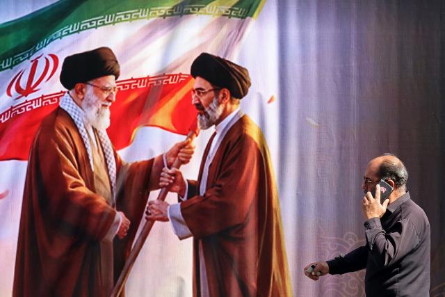 A man walks past a billboard featuring the images of Iran's slain supreme leader Ali Khamenei (L) and his son, the current supreme leader Mojtaba Khamenei, along the street in Tehran on July 3, 2026. Preparations for the funeral of Iran's slain supreme leader Ali Khamenei were in full swing on July 3, with authorities expecting millions and a coterie of foreign dignitaries to attend the official ceremony. (Photo by ATTA KENARE / AFP) / 