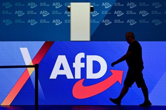 A worker walks past a screen displaying the logo of far-right Alternative for Germany (AfD) on the eve of the party congress at the Messe Erfurt fairground in Erfurt, eastern Germany on July 3, 2026, where the conference will be held from July 4-5. The AfD is holding its federal party conference in Erfurt this weekend to fill key leadership positions. Tens of thousands of people are expected to take part in protests against the gathering of the party. Several alliances have called for resistance, plans include colourful events and demonstrations, as well as blockades. A large police force has been deployed. (Photo by John MACDOUGALL / AFP)