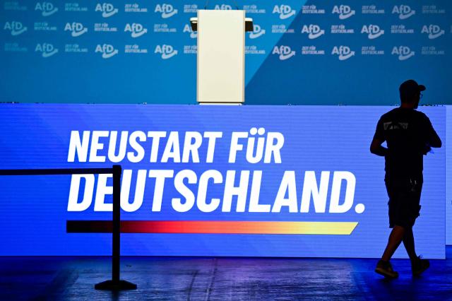 A worker walks past a screen displaying the slogan "New start for Germany" on the eve of the party congress of far-right Alternative for Germany (AfD) at the Messe Erfurt fairground in Erfurt, eastern Germany on July 3, 2026, where the conference will be held from July 4-5. The AfD is holding its federal party conference in Erfurt this weekend to fill key leadership positions. Tens of thousands of people are expected to take part in protests against the gathering of the party. Several alliances have called for resistance, plans include colourful events and demonstrations, as well as blockades. A large police force has been deployed. (Photo by John MACDOUGALL / AFP)