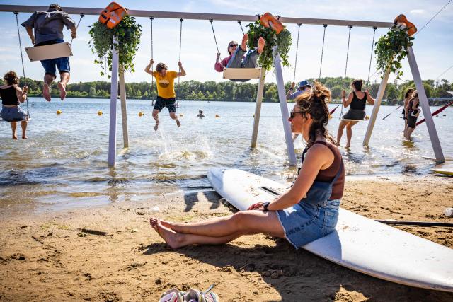 A woman sits on a surfboard as festival-goers swing on swings on the shore of the Groene Heuvels lake during the first day of the Down the Rabbit Hole music festival in Beuningen, eastern Netherlands on July 3, 2026. (Photo by Marcel Krijgsman / ANP / AFP) / Netherlands OUT