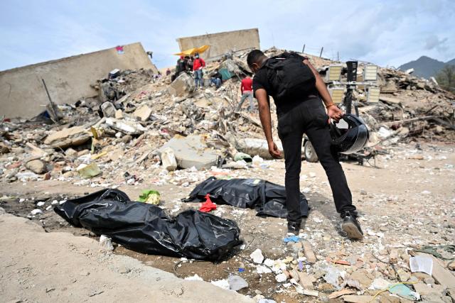 A man leans over body bags placed on the ground to read identification tags during recovery operations amid the rubble of collapsed buildings in Caraballeda, La Guaira state, Venezuela, on July 3, 2026. The death toll from Venezuela's twin earthquakes has risen to 2,595, Interim President Delcy Rodriguez said on July 2. (Photo by MARTIN BERNETTI / AFP)
