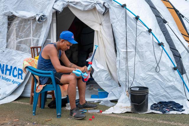 A displaced man repairs a scooter for his son outside a tent at the Cesar Nieves baseball stadium in the La Angustia neighborhood of Catia La Mar, La Guaira state, Venezuela, on July 3, 2026, where displaced people have taken refuge. The death toll from Venezuela's devastating twin earthquakes has risen to 2,645, with more than 12,600 injured, according to new official figures released on July 3. (Photo by Miguel MEDINA / AFP)