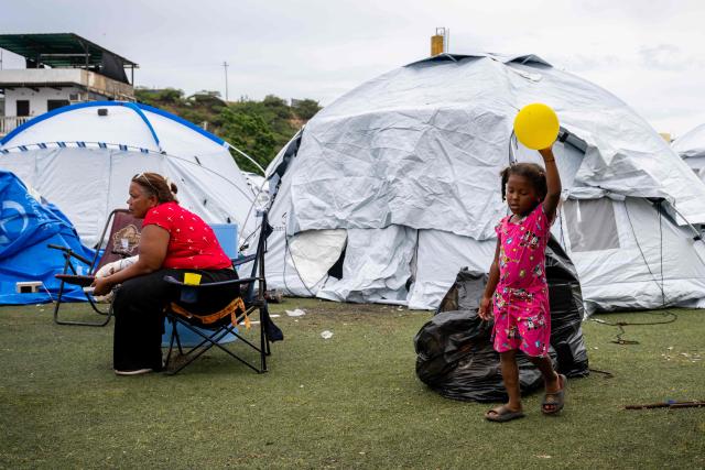 A child holds a yellow balloon as a displaced woman sits near tents at the Cesar Nieves baseball stadium in the La Angustia neighborhood of Catia La Mar, La Guaira state, Venezuela, on July 3, 2026, where displaced people have taken refuge. The death toll from Venezuela's devastating twin earthquakes has risen to 2,645, with more than 12,600 injured, according to new official figures released on July 3. (Photo by Miguel MEDINA / AFP)