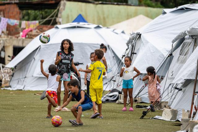 Children play with a ball near tents at the Cesar Nieves baseball stadium in the La Angustia neighborhood of Catia La Mar, La Guaira state, Venezuela, on July 3, 2026, where displaced people have taken refuge. The death toll from Venezuela's devastating twin earthquakes has risen to 2,645, with more than 12,600 injured, according to new official figures released on July 3. (Photo by Miguel MEDINA / AFP)