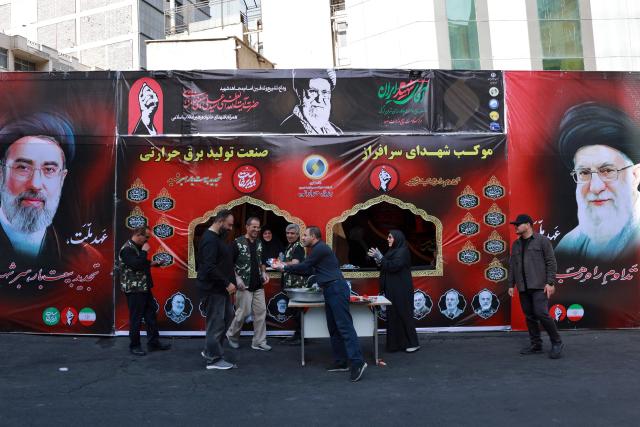 A volunteer hands out refreshments in front of portraits of Iran's supreme leader Ayatollah Mojtaba Khamenei (L) and slain supreme leader Ayatollah Ali Khamenei (R) while mourners make their way to the Grand Mosalla to pay their final respects at the start of his funeral ceremonies in Tehran on July 4, 2026. Vast crowds of Iranians loyal to the Islamic republic massed in Tehran on July 4 to begin almost a week of funeral ceremonies for slain supreme leader Ali Khamenei, which authorities want to send a message of defiance to the West after the war with the United States and Israel. (Photo by AFP) / 