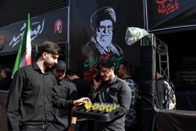A volunteer hands out refreshments in front of portraits of slain supreme leader Ayatollah Ali Khamenei (R) while mourners make their way to the Grand Mosalla to pay their final respects at the start of his funeral ceremonies in Tehran on July 4, 2026. Vast crowds of Iranians loyal to the Islamic republic massed in Tehran on July 4 to begin almost a week of funeral ceremonies for slain supreme leader Ali Khamenei, which authorities want to send a message of defiance to the West after the war with the United States and Israel. (Photo by AFP) / 