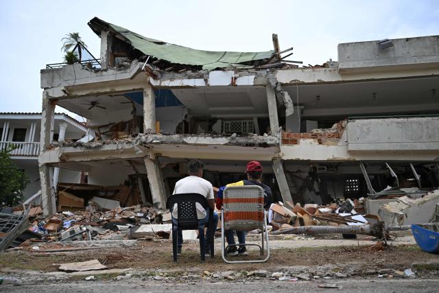 EDITORS NOTE: Graphic content / People sit in front of a destroyed building in La Guaira, Venezuela on July 4, 2026, following the June 24 twin earthquakes. With little hope of finding loved ones alive, frustrated families of those affected by Venezuela's double earthquakes clamor for help to recover their loved one's bodies as uncertainty grows over the death toll from the tragedy. (Photo by Raul ARBOLEDA / AFP)