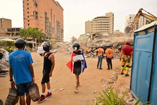 Residents carry supplies past a collapsed building as rescue workers continue operations in Catia La Mar, La Guaira state, on the outskirts of Caracas, on July 4, 2026, following the June 24 twin earthquakes. The death toll from Venezuela's devastating twin earthquakes has risen to 2,954, according to official figures released on Saturday. (Photo by MARTIN BERNETTI / AFP)