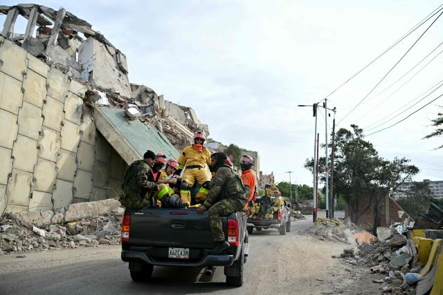 Brazilian rescuers ride in the back of pickup trucks past collapsed buildings as they head to carry out search and rescue operations in Catia La Mar, La Guaira state, on the outskirts of Caracas, on July 4, 2026, following the June 24 twin earthquakes. The death toll from Venezuela's devastating twin earthquakes has risen to 2,954, according to official figures released on Saturday. (Photo by MARTIN BERNETTI / AFP)