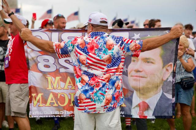 A man wears a shirt featuring images of US President Donald Trump and holds up a banner featuring Secretary of State Marco Rubio as people gather near the Washington Monument after being evacuated from the event site of the "Salute to America" Independence Day celebration due to thunderstorms in the area in Washington, DC, on July 4, 2026. The nation is marking the 250th anniversary of the United States Declaration of Indpendence. (Photo by Amid FARAHI / AFP)