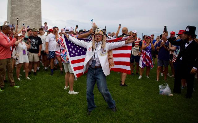A person wearing a feathered headdress holds a US flag as attendees gather near the Washington Monument after being evacuated from the event site of the "Salute to America" Independence Day celebration due to thunderstorms in the area in Washington, DC, on July 4, 2026. The nation is marking the 250th anniversary of the United States Declaration of Indpendence. (Photo by Amid FARAHI / AFP)
