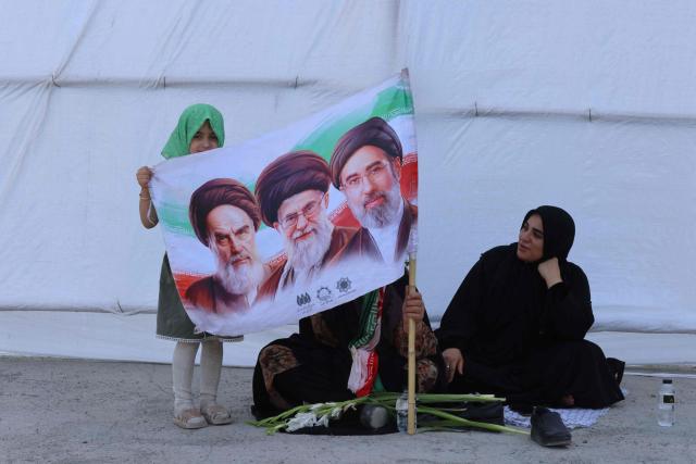 Mourners hold a flag showing the late founder of the Islamic Revolution supreme leader Ayatollah Ruhollah Khomeini (L), the Iran's slain supreme leader Ali Khamenei (C) and his son, the current supreme leader Mojtaba Khamenei as they gather to pray for Ali Khamenei during the second day of funeral ceremonies at the Grand Mosalla in Tehran on July 5, 2026. Prayers were held July 5 over the casket of Iran's late supreme leader Ali Khamenei, the second day of funeral ceremonies that have drawn huge crowds to pay their final respects in Tehran. Khamenei ruled the Islamic republic from 1989 until he was killed aged 86 in an airstrike on the first day of the US-Israeli war with Iran on February 28. (Photo by ATTA KENARE / AFP) / 