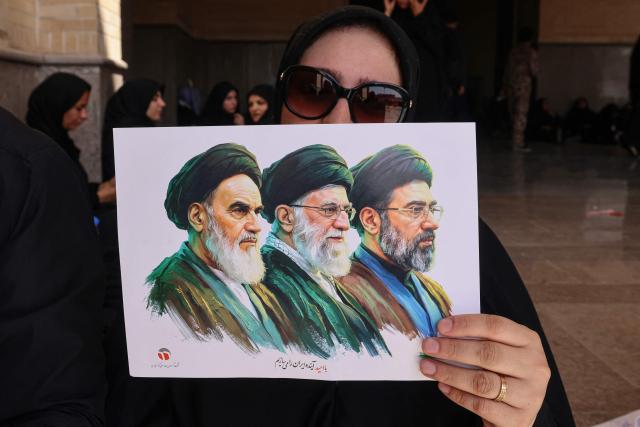 A mourner holds a picture showing the late founder of the Islamic Revolution supreme leader Ayatollah Ruhollah Khomeini (L), Iran's slain supreme leader Ali Khamenei (C) and his son, the current supreme leader Mojtaba Khamenei, as she pays her respect for Ali Khamenei at the Grand Mosalla during the second day of his funeral ceremonies in Tehran on July 5, 2026. Prayers were held July 5 over the casket of Iran's late supreme leader Ali Khamenei, the second day of funeral ceremonies that have drawn huge crowds to pay their final respects in Tehran. Khamenei ruled the Islamic republic from 1989 until he was killed aged 86 in an airstrike on the first day of the US-Israeli war with Iran on February 28. (Photo by ATTA KENARE / AFP) / 