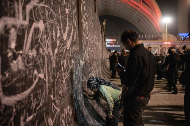 A girl writes a condolence message with chalk on a wall alongside other inscriptions as people gather to mourn Iran's slain supreme leader Ayatollah Ali Khamenei during the funeral ceremonies at the Grand Mosalla in Tehran on July 5, 2026. Iranian leaders on July 5 joined prayers over the casket of late supreme leader Ali Khamenei during a second day of funeral ceremonies, which was declared a public holiday across Iran. Khamenei's body will be moved out of the Grand Mosalla later in the day, where it is lying in state, as part of preparations for processions through the capital the following day. (Photo by AFP) / 