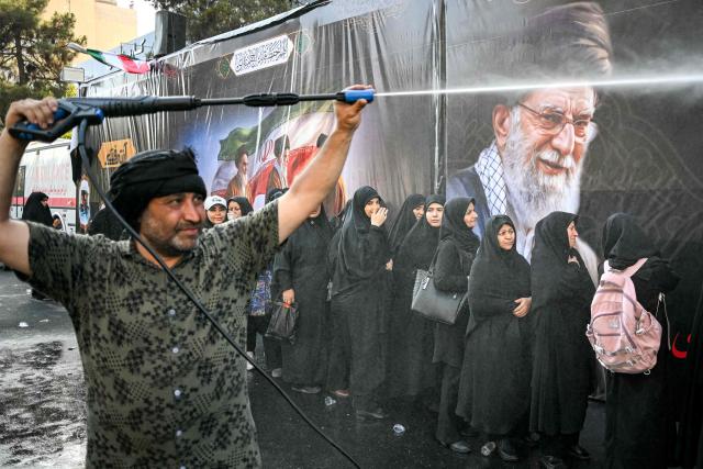 People are sprayed with water to cool off during the heat while queueing for food on their way to the Grand Mosalla in Tehran on July 5, 2026, for the funeral ceremonies of Iran's slain supreme leader Ayatollah Ali Khamenei. Iranian leaders on July 5 joined prayers over the casket of late supreme leader Ali Khamenei during a second day of funeral ceremonies, which was declared a public holiday across Iran. Khamenei's body will be moved out of the Grand Mosalla later in the day, where it is lying in state, as part of preparations for processions through the capital the following day. (Photo by Ozan KOSE / AFP) / 