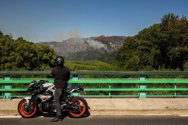 A motorcyclist stops on a road to watch the fire flare up again on a hill in the town of Ille sur Tet as a wildfire rages in the southern France department of Pyrenees-Orientales where 700 hundred firefighters backed by special aircraft battled to control a "gigantic" blaze spreading in a hard-to-reach remote area, with more than 10,000 local residents evacuated, near Perpignan on July 6, 2026. Wildfires raged across southern Europe, forcing thousands of people to evacuate their homes and prompting officials to ban spectators from a stage of the storied Tour de France cycling race. (Photo by Jean-Christophe MILHET / AFP)