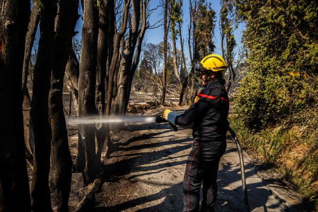 A firefighter sprays water on smoke plumes to prevent the fire from reigniting in the town of Ille sur Tet as a wildfire rages in the southern France department of Pyrenees-Orientales where 700 hundred firefighters backed by special aircraft battled to control a "gigantic" blaze spreading in a hard-to-reach remote area, with more than 10,000 local residents evacuated, near Perpignan on July 6, 2026. Wildfires raged across southern Europe, forcing thousands of people to evacuate their homes and prompting officials to ban spectators from a stage of the storied Tour de France cycling race. (Photo by Jean-Christophe MILHET / AFP)