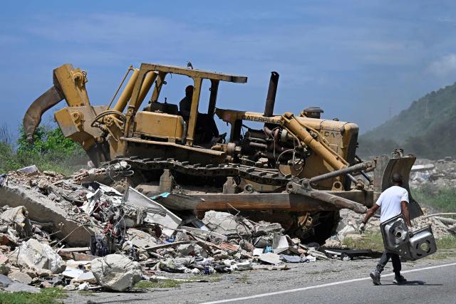 Heavy machinery clears debris from collapsed buildings as a person carries salvaged belongings along a roadside in the Caribe sector of Caraballeda, La Guaira state, Venezuela, on July 6, 2026, following powerful earthquakes that struck the regio. The earthquakes that struck Venezuela on June 24 have claimed the lives of at least 3,342 people, the Venezuelan government reported on July 5. (Photo by Federico PARRA / AFP)