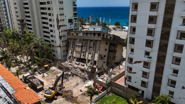 This aerial picture shows heavy machinery working on a residential building partially sunk into the ground near Playa Escondida in Tanaguarena, La Guaira state, Venezuela, on July 8, 2026. Venezuela's government called on July 8, 2026 for the release of frozen assets to help raise funds for the country's recovery from last month's twin earthquakes that killed at least 3,600 people. (Photo by Miguel MEDINA / AFP)