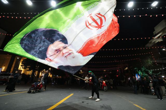 A man waves a flag near Imam Reza's shrine as mourners arrive for the funeral of slain Iranian leader Ali Khamenei and members of his family in Mashhad early on July 9, 2026. Iranians were preparing on July 9 to bury their slain leader Ali Khamenei in his hometown of Mashhad in northeast Iran, as new US strikes threatened to trigger a fresh escalation in the Middle East war. The late supreme leader's burial follows a multi-day funeral ceremony that attracted millions of mourners across various cities in both Iran and Iraq. (Photo by ATTA KENARE / AFP) / 