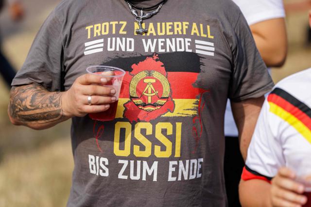 A participant wears a shirt reading "despite the fall of the Wall and the political turnaround- Ossi to the end" during an election campaign event of the far-right Alternative for Germany (AfD) party's and a motorbike and moped ride in Jessnitz, eastern Germany on June 28, 2026. Fans of far-right Alternative for Germany party's top candidate for the regional elections in Saxony-Anhalt, Ulrich Siegmund joined him for a countryside ride on Simson-brand scooters, that have become a symbol of East German identity. Descendants of the Simson firm's namesake Jewish founders, who gave up the company to escape persecution in Nazi Germany, have denounced the AfD's appropriation of the brand name. (Photo by Jens SCHLÜTER / AFP)