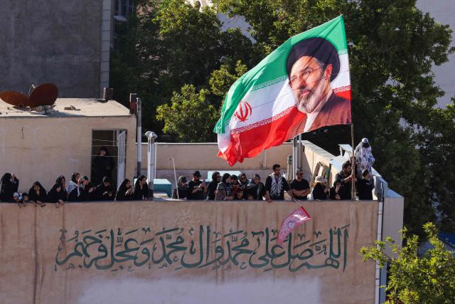 A flag of Iran bearing the image of Iran's current supreme leader Mojtaba Khamenei is seen on top of a building as people watch mourners take part in a funeral procession for Iran's slain supreme leader Ali Khamenei and members of his family, before he is buried at the Shrine of Imam Reza, Iran's most revered place of worship, in Mashhad on July 9, 2026. Braving sweltering heat and calling for revenge, Iranians loyal to the Islamic republic massed on July 9 for the burial of slain supreme leader Ali Khamenei in his hometown of Mashhad. (Photo by ATTA KENARE / AFP) / 