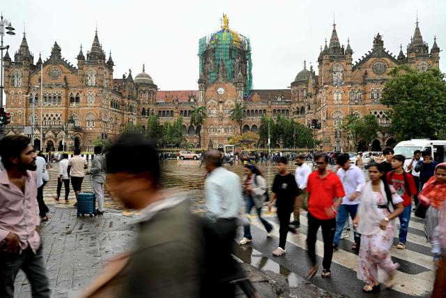 This photograph taken on July 7, 2026 shows pedestrians crossing a street to walk onto a footpath near Chhatrapati Shivaji Maharaj Terminus (CSMT) railway station in Mumbai. In India, a pavement can be many things -- hawking site, parking area, garbage dumping space -- but rarely what it is meant to be: a safe unobstructed path to walk on. (Photo by Indranil Mukherjee / AFP)