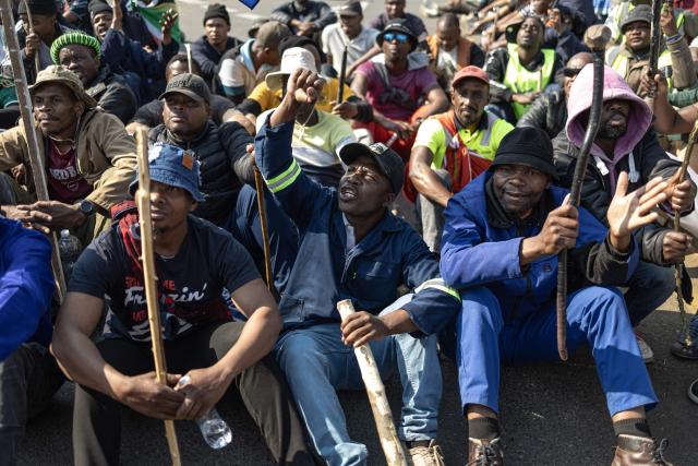 A protester gestures as others gather in front a Builders hardware store as South African Police Service (SAPS) officers stand guard during an anti-migration demonstration in Norwood on July 11, 2026. Protests that began with groups like March and March have since spread to ordinary residents taking to the streets, as anti-migrant sentiment continues to spread across South Africa. (Photo by EMMANUEL CROSET / AFP)