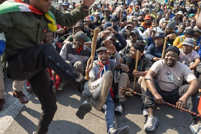Protesters dance as others gather in front a Builders hardware store as South African Police Service (SAPS) officers stand guard during an anti-migration demonstration in Norwood on July 11, 2026. Protests that began with groups like March and March have since spread to ordinary residents taking to the streets, as anti-migrant sentiment continues to spread across South Africa. (Photo by EMMANUEL CROSET / AFP)
