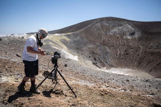 A vulcanologist from the University of Munich works with a laser instrument to gather data from a drone and collect data from gas emissions in the crater of La Fossa, on Vulcano Island, one of the seven islands of the Aeolian Archipelago of Sicily, on July 9, 2026. A team of researchers from Germany are testing a new system for measuring volcanic gases to better anticipate future eruptions. Hovering over the crater of La Fossa, a drone positions itself in front of a laser beam set at the edge of the chasm on Vulcano, off Sicily, and collect data. (Photo by Marco Bertorello / AFP)