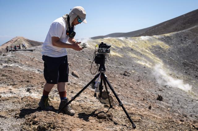 A vulcanologist from the University of Munich works with a laser instrument to gather data from a drone and collect data from gas emissions in the crater of La Fossa, on Vulcano Island, one of the seven islands of the Aeolian Archipelago of Sicily, on July 9, 2026. A team of researchers from Germany are testing a new system for measuring volcanic gases to better anticipate future eruptions. Hovering over the crater of La Fossa, a drone positions itself in front of a laser beam set at the edge of the chasm on Vulcano, off Sicily, and collect data. (Photo by Marco Bertorello / AFP)