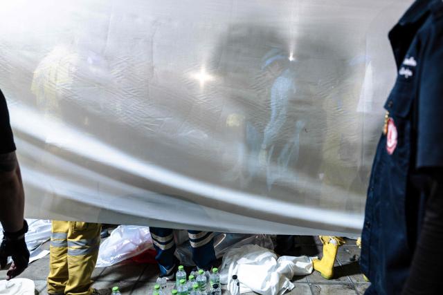 Emergency workers stand next to a white fabric displayed at the scene after a massive fire engulfed a bar in Bangkok, early on July 13, 2026. A fire in a bar in the Thai capital Bangkok killed at least 27 people late Sunday night, Prime Minister Anutin Charnvirakul told local media. (Photo by chanakarn LAOSARAKHAM / AFP)