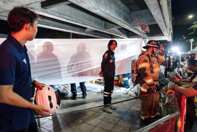 Emergency workers stand next to a white fabric displayed at the scene after a massive fire engulfed a bar in Bangkok, early on July 13, 2026. A fire in a bar in the Thai capital Bangkok killed at least 27 people late Sunday night, Prime Minister Anutin Charnvirakul told local media. (Photo by chanakarn LAOSARAKHAM / AFP)
